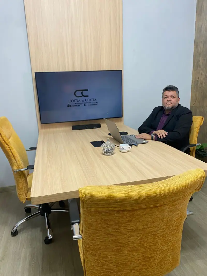Sala de reuniões do escritório com mesa de madeira, cadeiras amarelas e o Doutor Jocinei Costa ao fundo